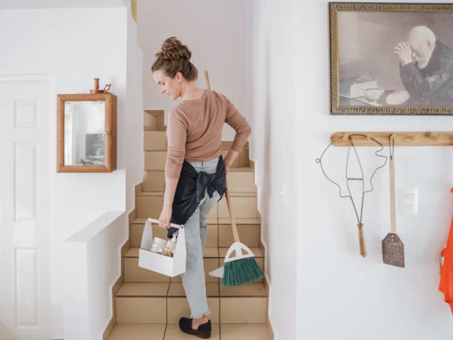 Cleaner carrying a caddy and broom up the stairs of a home